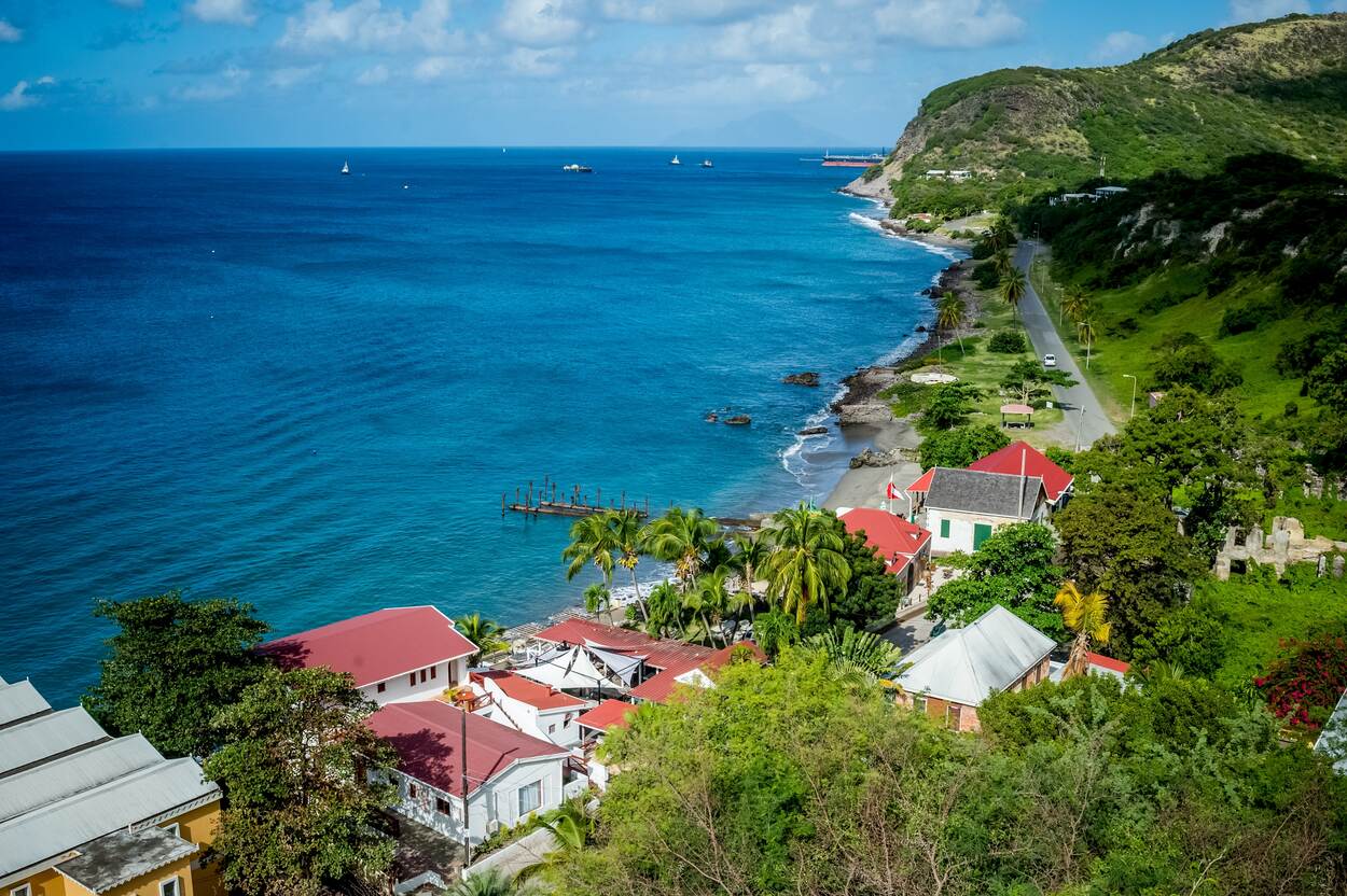 Een stuk kustlijn van het eiland Sint Eustatius gezien vanaf een heuvel. Links de zee met enkele boten. Rechts het heuvelachtige en groen begroeide eiland. Op de voorgrond staan enkele huizen. Langs de kust loopt een geasfalteerde weg waar een witte auto overheen rijdt.