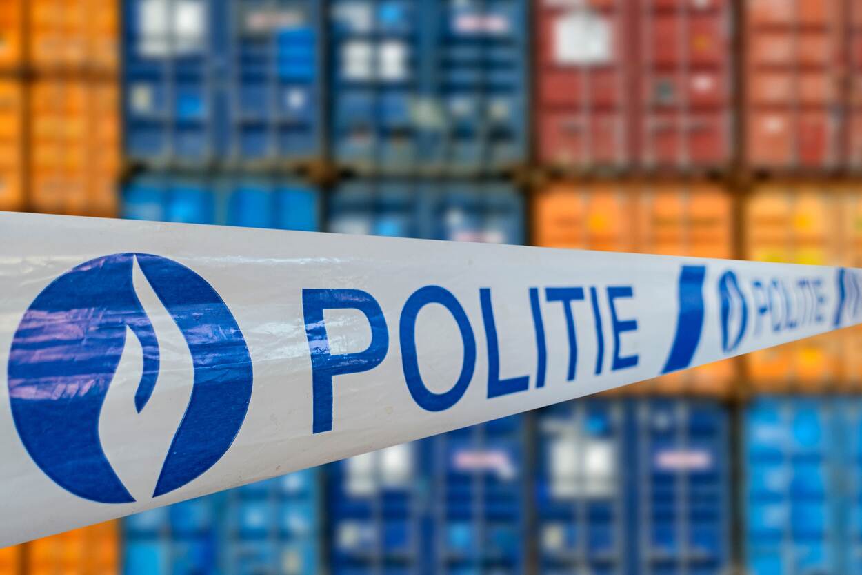Een wit afzetlint met in blauwe letters het woord politie en een blauw logo. Daarachter gestapelde zeecontainers in verschillende kleuren,