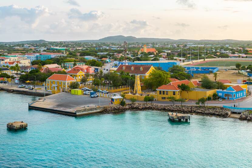 De cruisehaven van Bonaire vanaf zee gezien. Langs de kust loopt een geasfalteerde weg met daarlangs enkele parkeerplekken en een grote aanlegplaats. Direct achter de weg staan lage,  geel en blauw gekleurde gebouwen met een oranje dak.  Daarachter liggen een voetbalveld en nog meer gebouwen tot aan de heuvels op de horizon. Tussen de gebouwen staat veel groene begroeiiing.