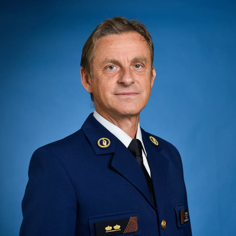 Portret van een man met kort bruin haar in een donkerblauw uniform met goudkleurige emblemen. De man kijkt recht in de camera en staat tegen een blauwe achtergrond..
